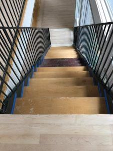 Staircase with light wood steps, dark metal railing, and blue painter's tape; viewed from above.
