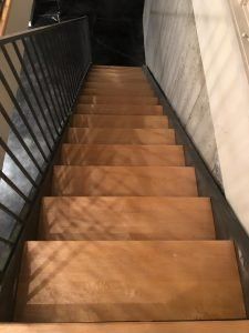 Wooden staircase descending into a dark space, with a metal railing on the left and a concrete wall on the right.