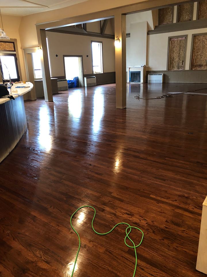 Shiny, dark hardwood floor in a large room with wooden support beams and a fireplace. A green cord is on the floor.